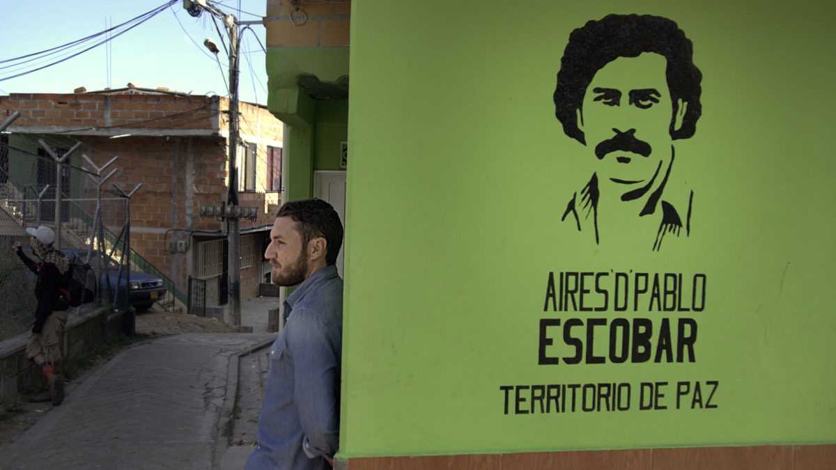 Doug Laux leans against a wall in the barrio with a stencil of Pablo Escobar?s face spray-painted on its surface.