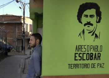 Doug Laux leans against a wall in the barrio with a stencil of Pablo Escobar?s face spray-painted on its surface.